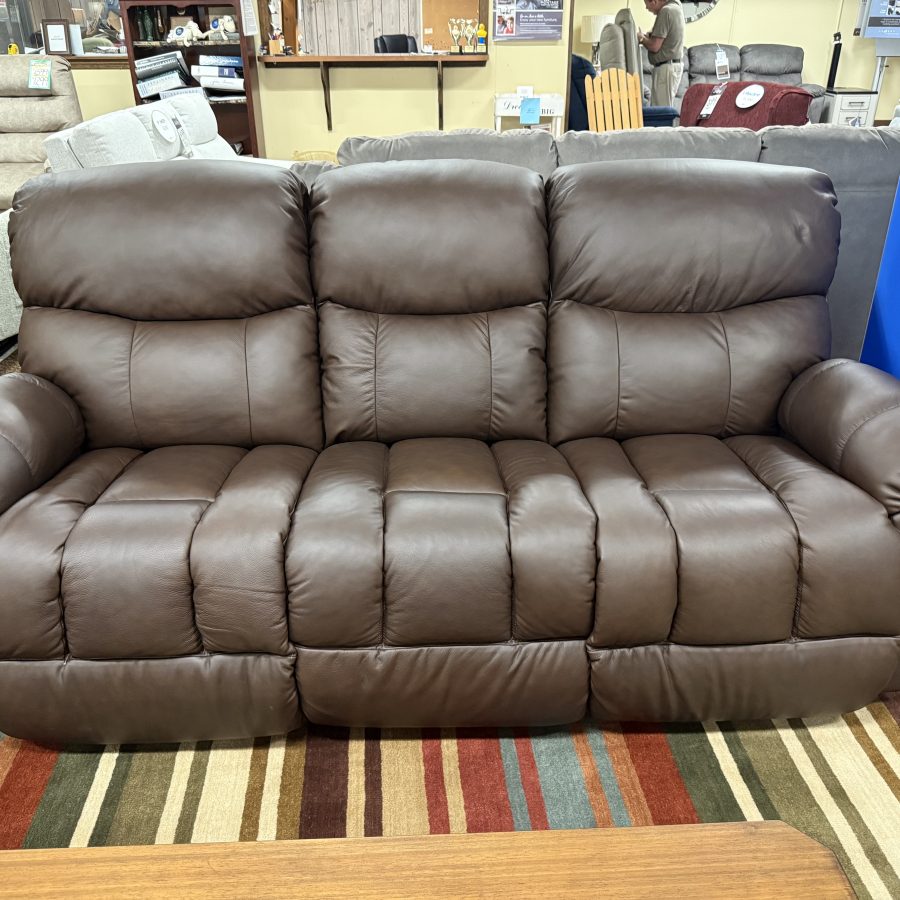 Brown leather reclining sofa with three back cushions on a striped showroom rug; price tag visible on the left.