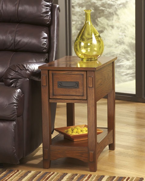 Oak wood side table with a small drawer and lower shelf, topped by a yellow glass vase beside a leather chair in a living room.